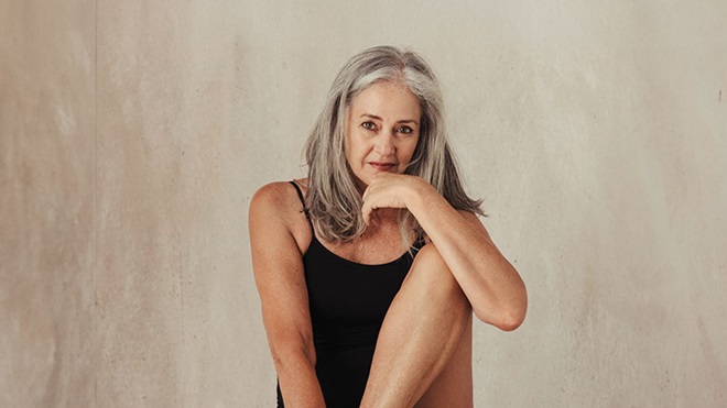 Senhora com cabelos compridos brancos e top interior preto, está sentada num banco e olha para a câmara com ar sério, fundo creme atrás