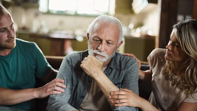 Homem com barbas e cabelo branco a sentir-se triste com um casal ao lado a consolá-lo - alusão ao tema do luto