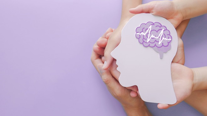 Mãos a segurar um recorte em forma de cabeça com um desenho de um cérebro e uma linha da vida; fundo lilás