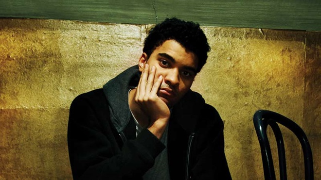 Grande plano de um jovem com ar deprimido, sentado numa cadeira, fundo amarelo escuro atrás 