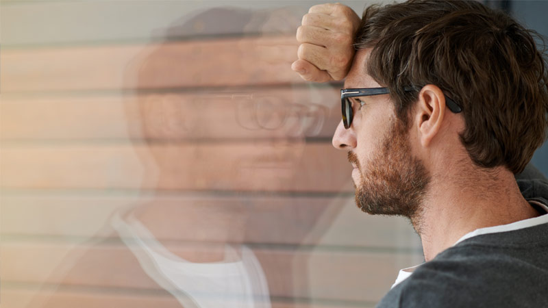 Grande plano de homem adulto com barba e óculos, encostado a uma janela com ar pensativo - alusão a saúde mental