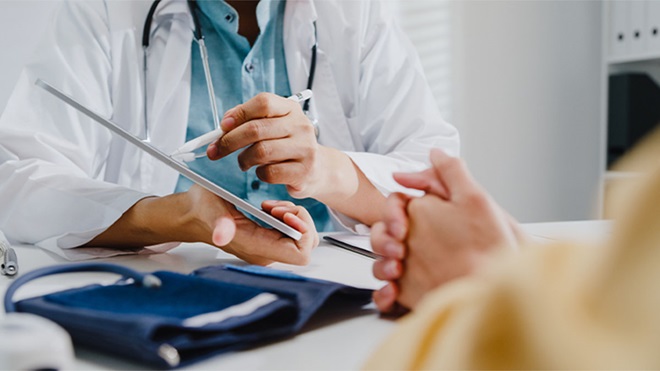 Profissional de saúde de bata branca escreve num formulário enquanto outra pessoa tem a mão apoiada na mesa; estetoscópio e braçadeira de tensão arterial visíveis.