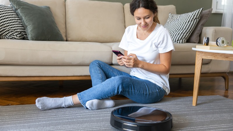 Mulher sentada no chão da sala põe o aspirador robot a funcionar.