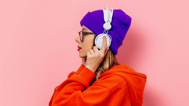 Jovem a ouvir música com auscultadores, com hoodie laranja e gorro roxo, num fundo rosa
