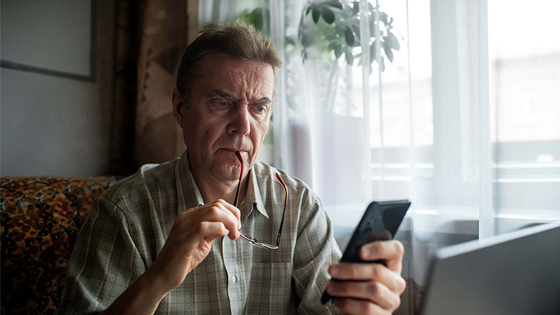 Pessoa sentada à mesa segura uns óculos e usa um smartphone, com computador portátil aberto à frente e luz natural a entrar pela janela.