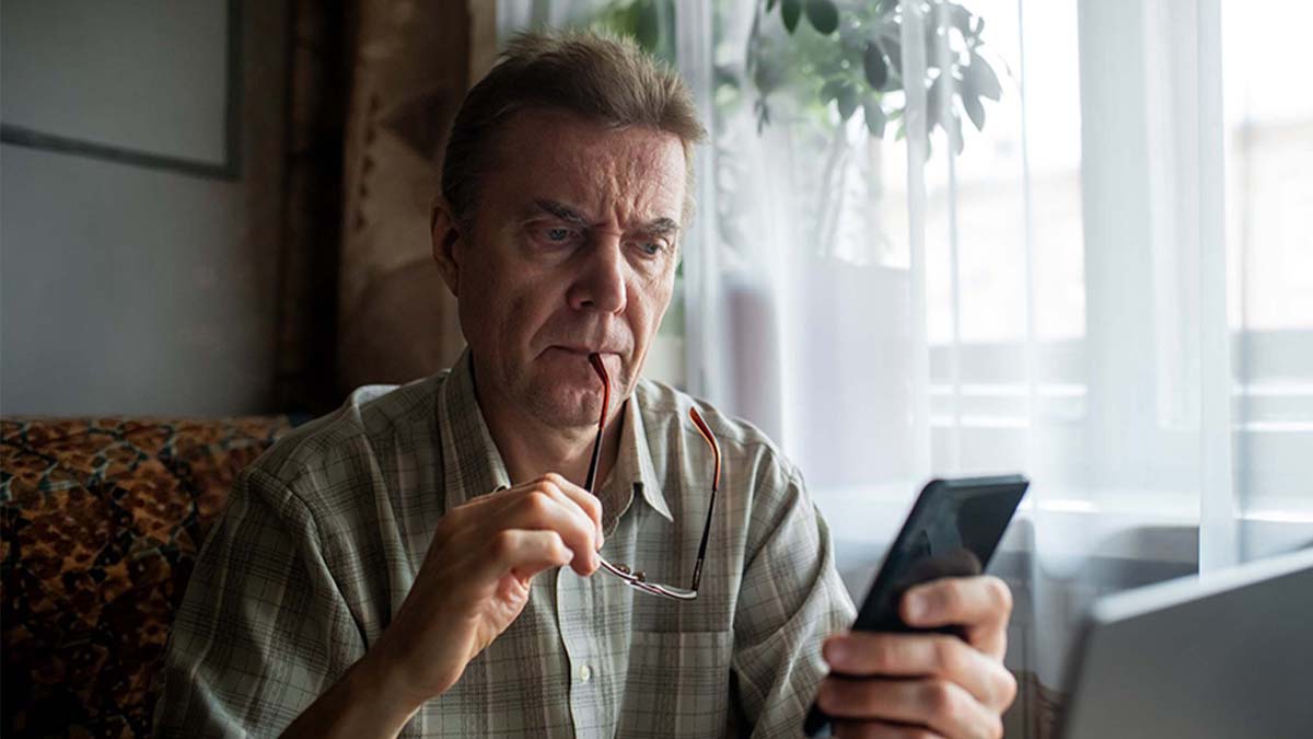 Pessoa sentada à mesa segura uns óculos e usa um smartphone, com computador portátil aberto à frente e luz natural a entrar pela janela.