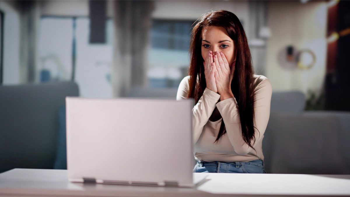 Mulher sentada numa secretária, com as mãos no rosto, em frente a um portátil aberto, num ambiente de escritório desfocado.