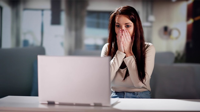 Mulher sentada numa secretária, com as mãos no rosto, em frente a um portátil aberto, num ambiente de escritório desfocado.