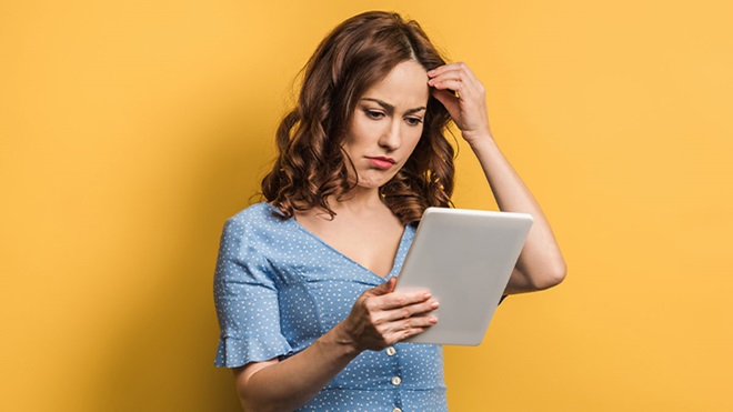 Mulher jovem com ar chateado a olhar para um tablet, fundo amarelo