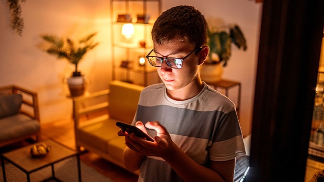 Adolescente num ambiente acolhedor de sala de estar, iluminado por luz quente, a segurar um telemóvel enquanto o utiliza, com plantas e prateleiras ao fundo.