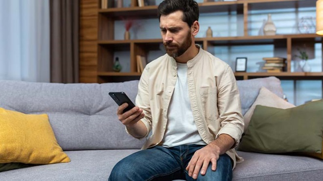 Pessoa sentada num sofá numa sala moderna, segurando um telemóvel e olhando para o ecrã com ar intrigado, com estante cheia de livros e objetos decorativos ao fundo.