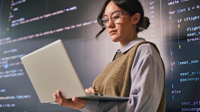 Pessoa com óculos usa portátil diante de ecrãs com linhas de código de programação.