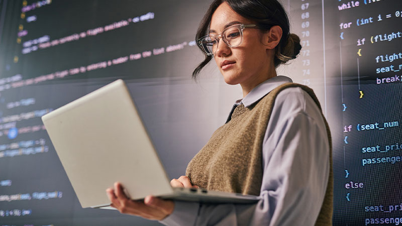 Pessoa com óculos usa portátil diante de ecrãs com linhas de código de programação.