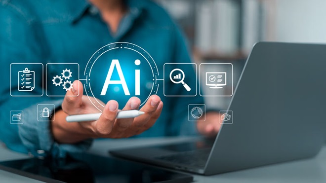 Grande plano de mão de homem com um símbolo sobre a imagem com as siglas "AI"; está sentado à mesa com um portátil e um tablet