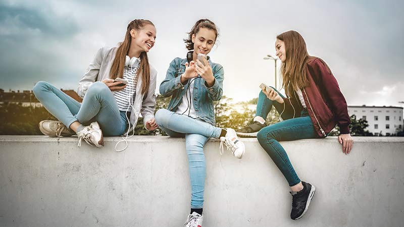 3 jovens sentadas em cima de um muro a ver os telemóveis