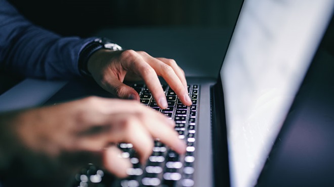 Grande plano de um computador portátil aberto, posicionado de lado para quem vê a imagem. Do lado esquerdo, sobre o teclado, estão duas mãos masculinas.