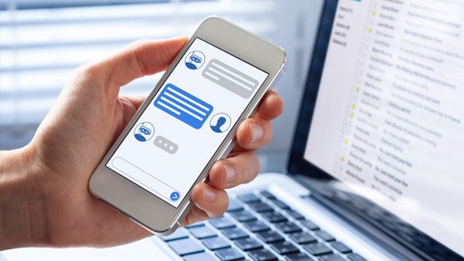 Mão segura um telemóvel com conversção com chatbot; atrás um portátil aberto
