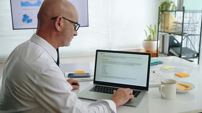 Homem careca de meia idade com óculos, vestido com camisa branca, a usar uma ferramenta de inteligência artificial à secretária num computador portátil