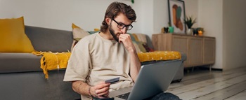 Homem a fazer compras online sentado no chão da sala, com ar preocupado, com um cartão de crédito na mão e um portátil no colo