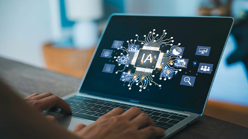 Mãos a escrever num portátil sobre uma mesa; no ecrã surge a sigla “IA” rodeada de ícones digitais interligados, sugerindo tecnologia e inteligência artificial.