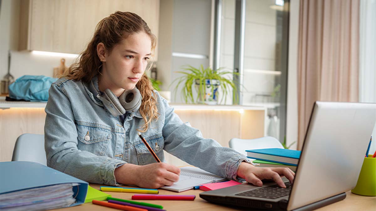 Rapariga jovem a estudar, sentada numa secretária num espaço luminoso com plantas, a olhar para o ecrã de um portátil enquanto escreve num caderno