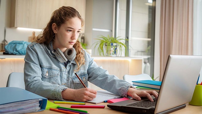 Rapariga jovem a estudar, sentada numa secretária num espaço luminoso com plantas, a olhar para o ecrã de um portátil enquanto escreve num caderno