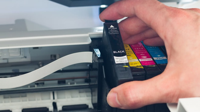 Plano apertado do interior de uma impressora, com uma mão a retirar um tinteiro de tinta preta do compartimento dos tinteiros