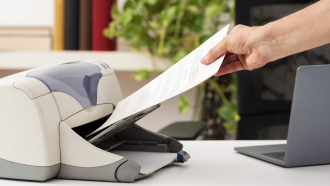 Mão de mulher a colocar folhas de papel branco numa impressora que está em cima de uma mesa com um portátil ao lado.