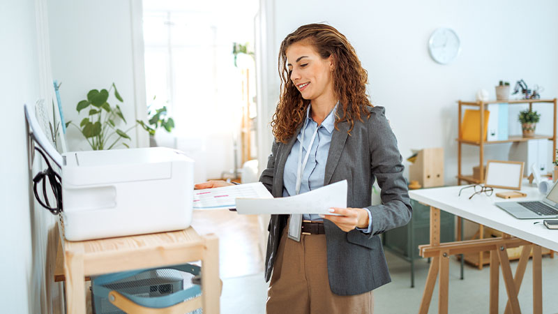 Pessoa num escritório luminoso segura papeis junto a uma impressora, com secretária, plantas e prateleiras de trabalho ao fundo.