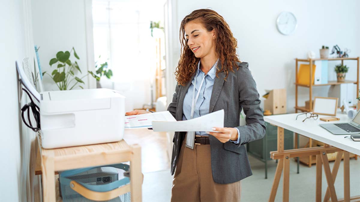 Pessoa num escritório luminoso segura papeis junto a uma impressora, com secretária, plantas e prateleiras de trabalho ao fundo.