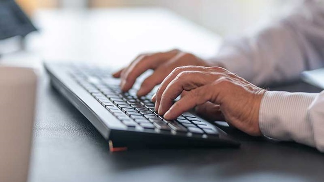 Mãos digitando sobre um teclado sem fio, instalado numa mesa escura
