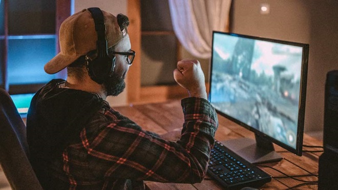 Homem com boné na cabeça e óculos, sentado numa cadeira a olhar para um ecrã onde joga um jogo de computador. Punho para cima mostrando vitória