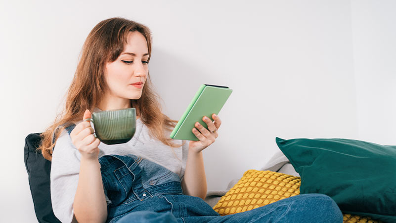 Mulher jovem sentada num sofá, com uma caneca numa mão e um ereader na outra, lê confortavelmente. 