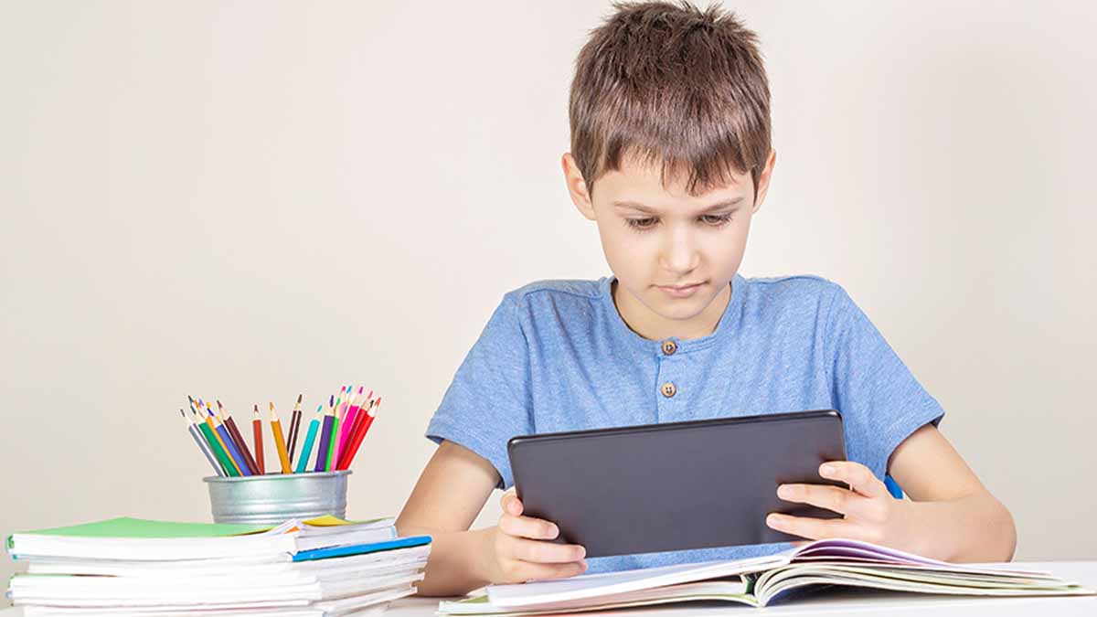 Rapaz com cerca de 9 anos segura num tablet sentado a uma mesa com material escolar