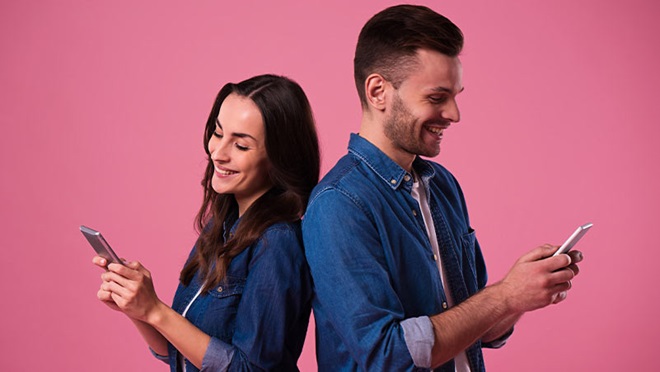 Um homem e uma mulher de costas voltadas, cada um com o seu telemóvel, sorriem para o ecrãn, fundo rosa. Alusão a apps de encontros