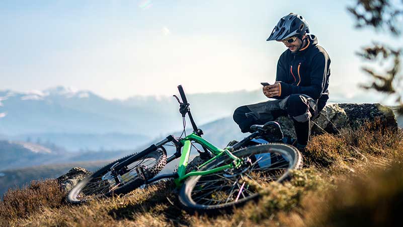 Atleta de downhill sentado no cimo da colina com a bicicleta ao lado, consulta um smartphone
