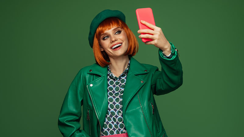 Mulher de gabardine e boina a tirar selfie com telemóvel, em fundo verde, foto de estúdio