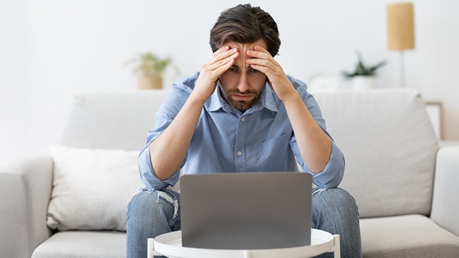 Homem adulto sentado no sofá da sala com um portátil à frente e com ar preocupado