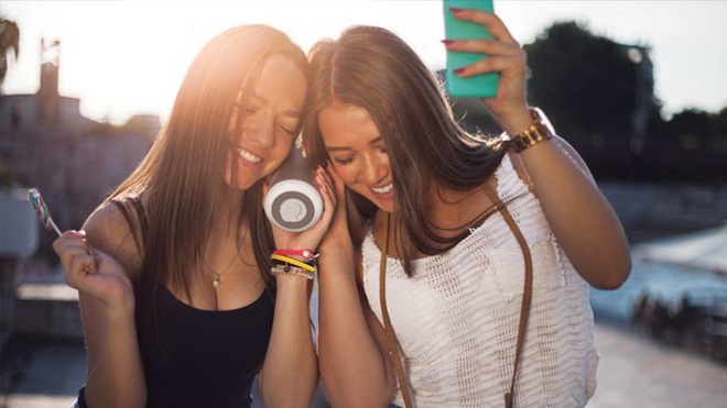 Duas jovens na rua, a ouvirem música através de uma coluna de som bluetooth emparelhada com o telemóvel
