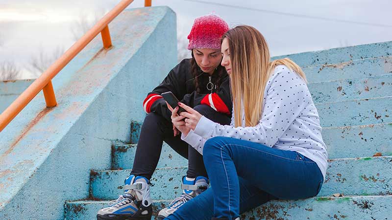 Duas jovens adolescentes, numa escadaria na rua, a olhar para um telemóvel