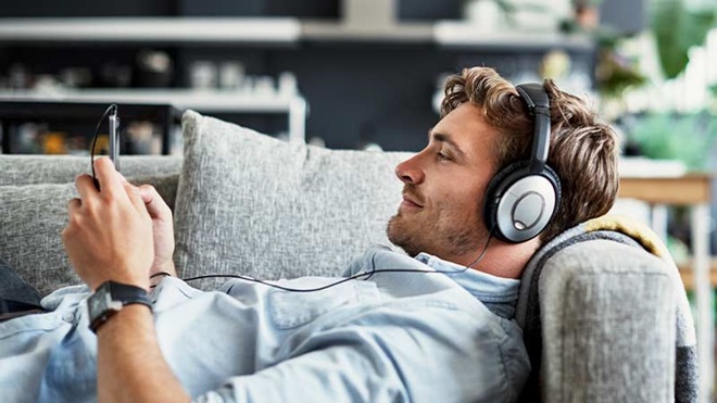 Homem deitado no sofá em casa, com phones, ouve música com o telemóvel