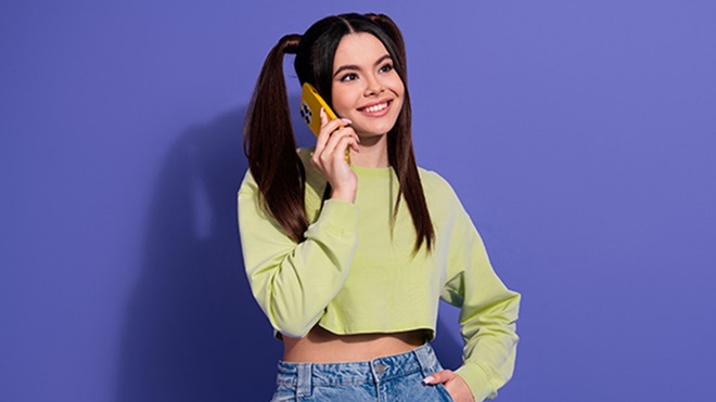 Jovem adulta morena e sorridente ao telemóvel, sobre fundo roxo 