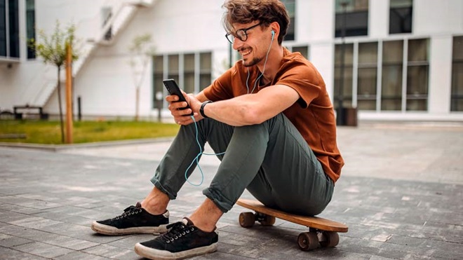 Home jovem sentado num skate, no exterior a consultar o telemóvel, com óculos e phones nos ouvidos