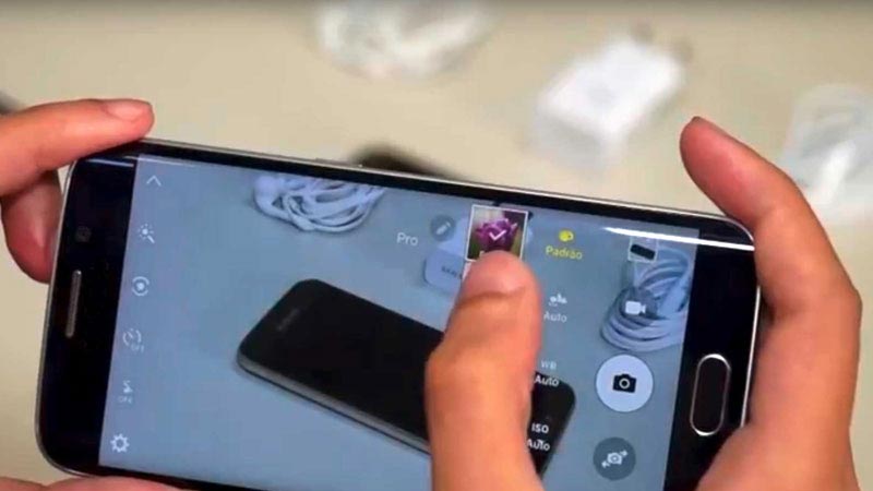Mãos a mexerem num smartphone para teste a telemóveis, com fichas brancas de carregadores atrás.