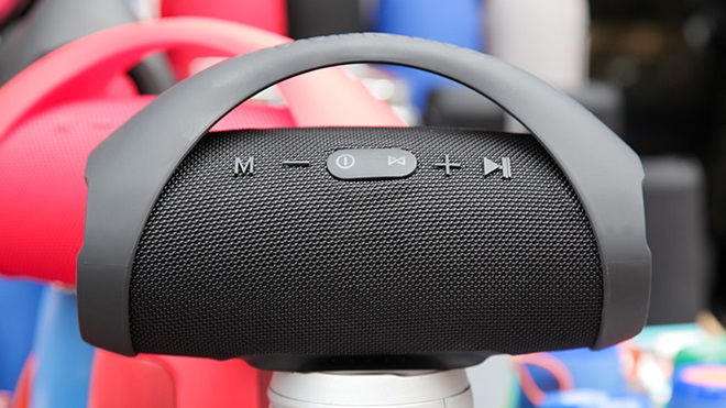 Em primeiro plano, uma coluna Bluetooth de cor preta e forma cilíndrica. No topo da coluna são visíveis os controlos para ligar, tocar música, avançar e aumentar e diminuir o som. A coluna possui uma pega rígida, em forma de meia lua, que começa nas pontas e contorna o topo da coluna, com os controlos. Por trás, só parcialmente visível, está uma coluna semelhante, mas de cor vermelha.