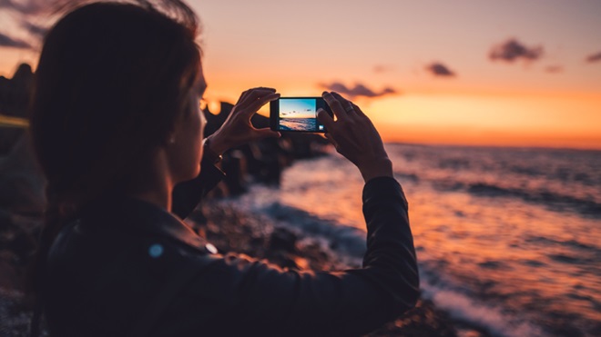 Mulher a fotografar com um telemóvel, junto ao mar, ao pôr-do-sol