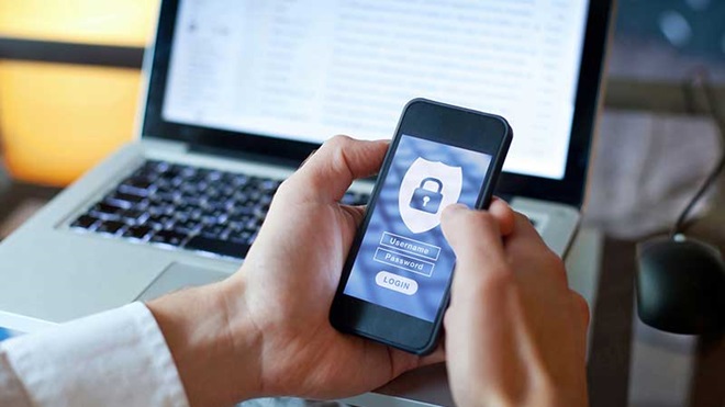 Homem ao computador com telemóvel nas mãos a mostrar um cadeado azul e branco no ecrã do smartphone.