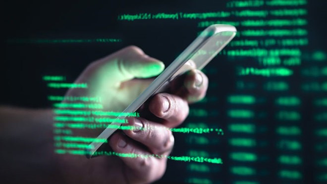 Imagem de uma mão segurando um telemóvel prateado, refletida num ecrã de fundo preto, com várias linhas de código de programação informática, em cor verde