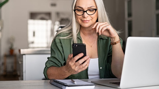 Senhora sentada à secretária segura no telemóvel; laptop na mesa (alusão a como transferir ficheiros do telemóvel para o computador)