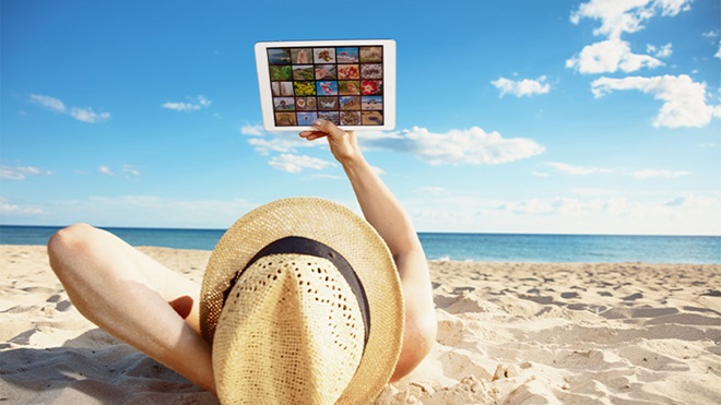 Pessoa deitada na areia da praia com chapéu de palha a ver TV no tablet